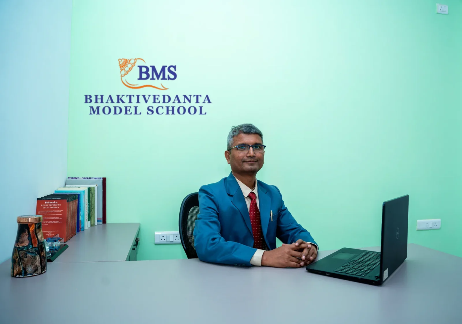 The Chairman of Bhaktivedanta Model School at his desk, the school's crest and name on the wall behind him