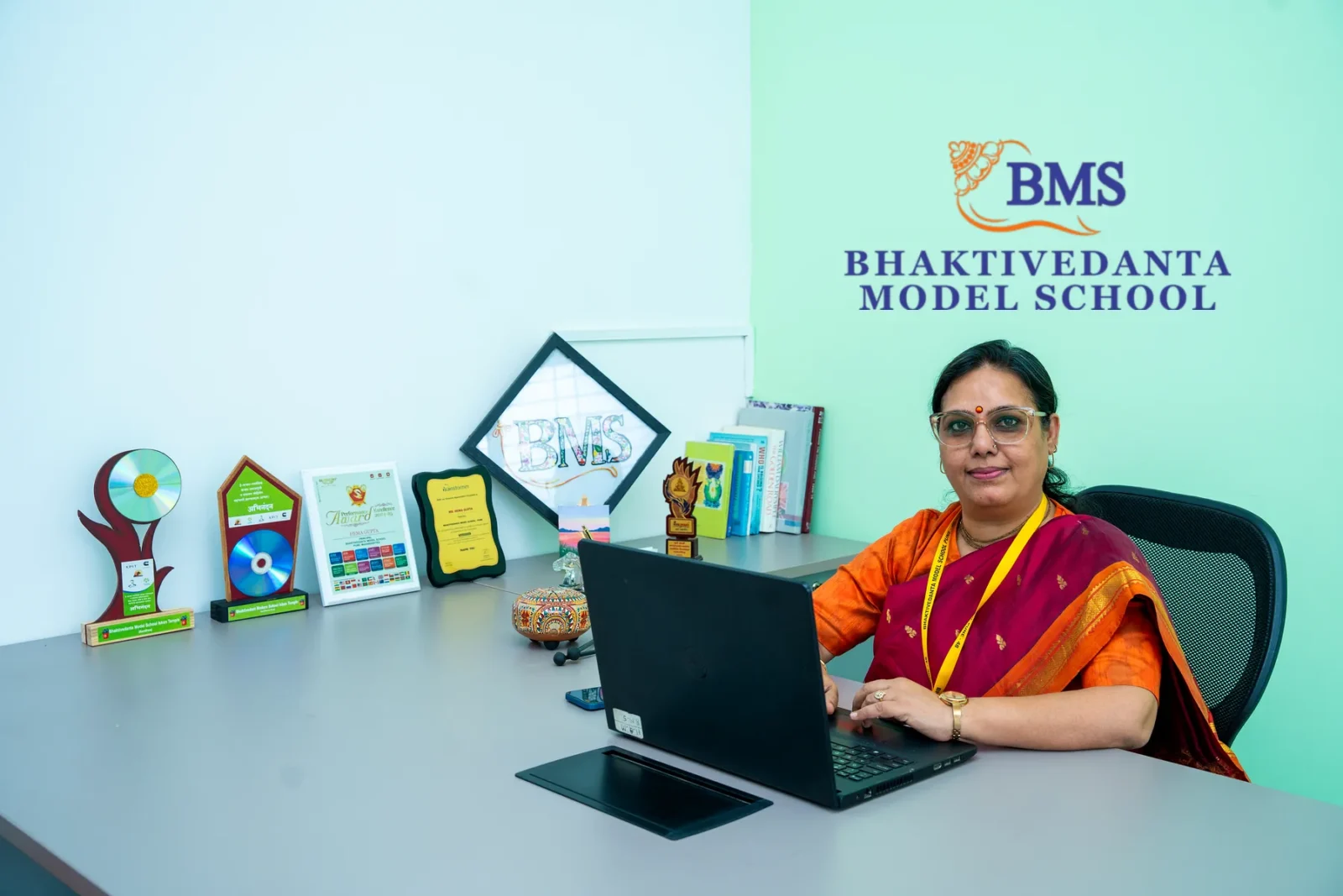 The Principal at work in the Principal's office, the school's crest and name on the wall behind her