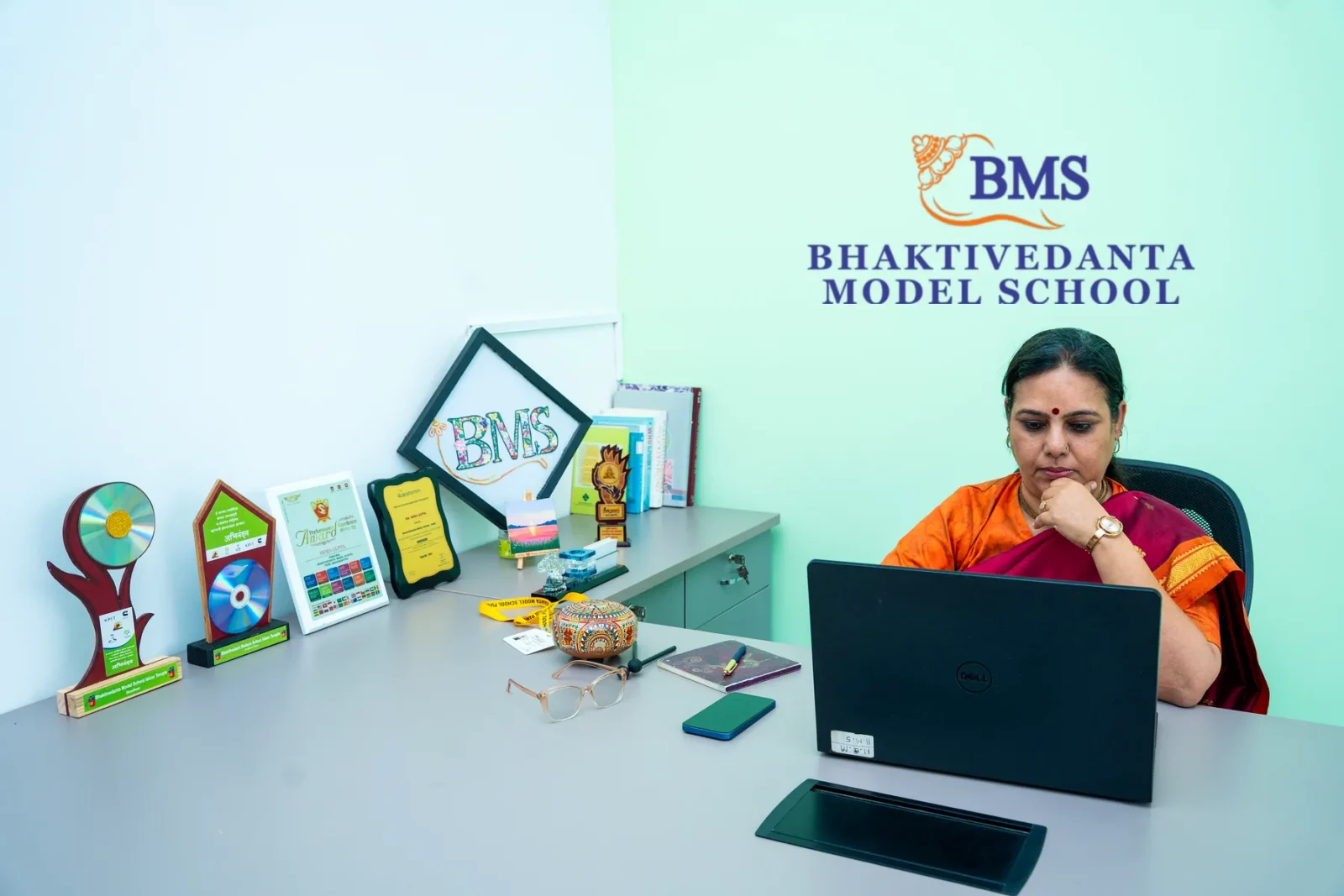 The Principal of Bhaktivedanta Model School at her desk, the school's crest and name on the wall behind her