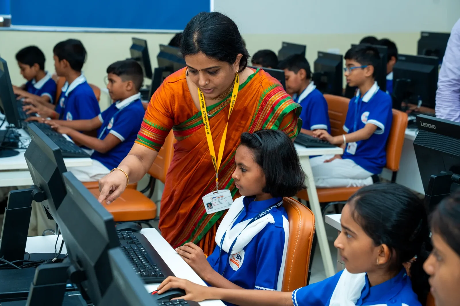 A teacher guiding a child at a workstation while the class works at the computers behind